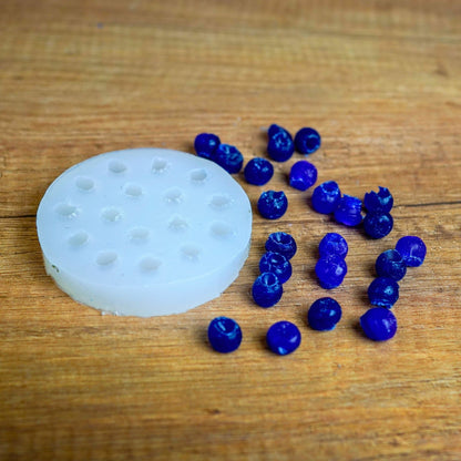 Runde weiße Silikonform mit Mini-Beeren-Mulden, daneben viele blau gegossene Wachsbeeren auf Holz