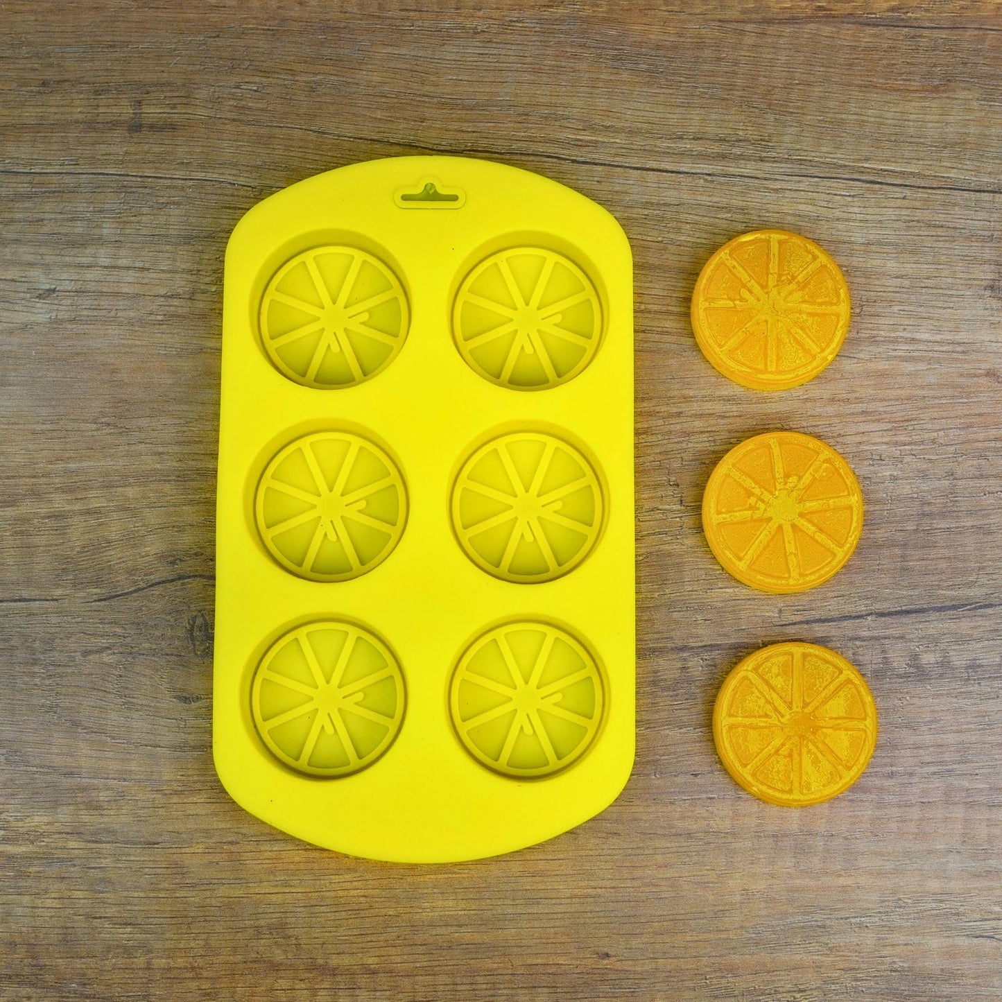 Gelbe Silikonform mit 6 Zitrusscheiben-Mulden, daneben drei fertig gegossene orange Fruchtscheiben